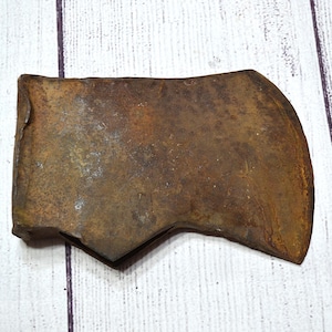 May include: A vintage axe head with a weathered, rusty appearance. The metal tool has a rectangular shape with a curved blade and a notched base. The axe head is a dark brown color and sits on a white wooden surface.
