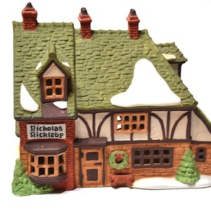 May include: A ceramic Christmas village house with a green shingled roof, snow accents, and a sign that reads "Nicholas Rickleb". The house features brown and white Tudor-style detailing, a wreath, and a small Christmas tree.
