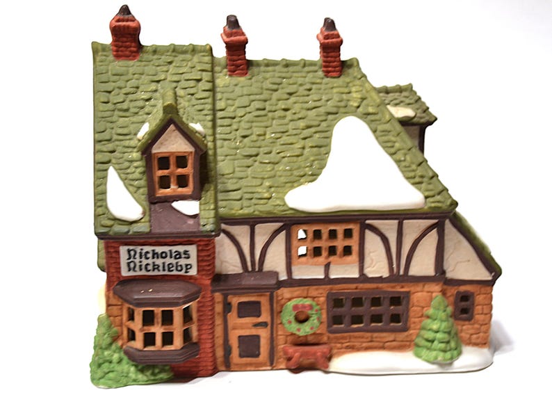 May include: A ceramic Christmas village house with a green shingled roof, snow accents, and a sign that reads "Nicholas Rickleb". The house features brown and white Tudor-style detailing, a wreath, and a small Christmas tree.