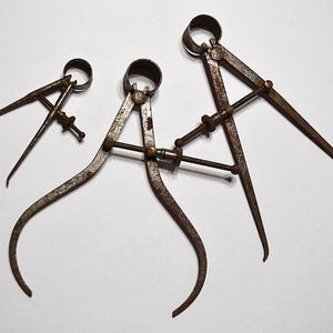 May include: Three antique rusty metal calipers of varying sizes are arranged on a white surface. The calipers have curved legs and adjustable screws, used for measuring distances. The tools are likely used for woodworking or metalworking.