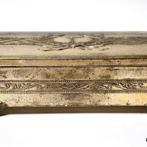 May include: A rectangular, antique metal box with a hinged lid. The box is silver-toned with ornate embossed designs, including a central emblem and floral patterns. The box rests on four decorative feet.