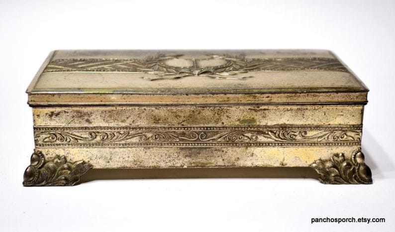 May include: A rectangular, antique metal box with a hinged lid. The box is silver-toned with ornate embossed designs, including a central emblem and floral patterns. The box rests on four decorative feet.