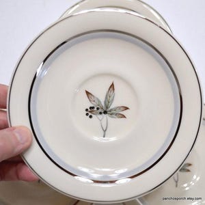 May include: Cream-colored saucer with a silver rim and a central leaf design. The leaf is depicted in shades of brown, green, and black. The saucer is part of a set, with several others visible in the background.