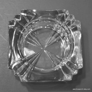 May include: Clear glass ashtray, square with a decorative, ridged rim. The interior has a circular, textured design with a starburst pattern. The ashtray's design is vintage and elegant, suitable for a classic aesthetic.
