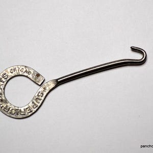 May include: A vintage metal shoe hook with a curved handle and a circular end with the words "SPATS CHICAGO SUPERIOR" embossed. The hook is silver-toned and appears to be made of steel.