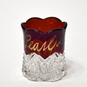 May include: A small, vintage glass with a ruby red interior and a clear, faceted base. The word "Pearls" is written in gold script on the red glass. The rim has a scalloped edge.