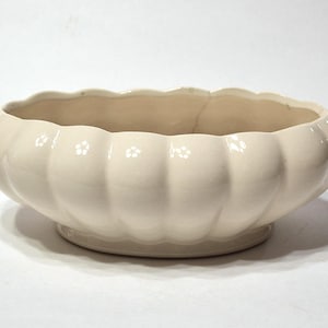 May include: Cream-colored ceramic bowl with a scalloped rim and a rounded, ribbed body. The bowl sits on a small, circular base. The interior of the bowl is a lighter shade of cream. A crack is visible on the rim.