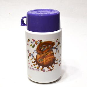 May include: A white insulated travel mug with a purple lid and handle. The mug features a graphic of a brown bag of Bertie Bott's Every Flavor Beans. The text "Bertie Bott's Flavor Beans" is printed on the mug.