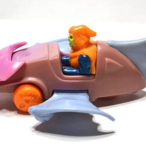 May include: A vintage toy vehicle featuring a character figure. The vehicle is primarily brown with pink and blue accents, and orange wheels. The character has an orange hood and green skin. The toy is on a white surface.