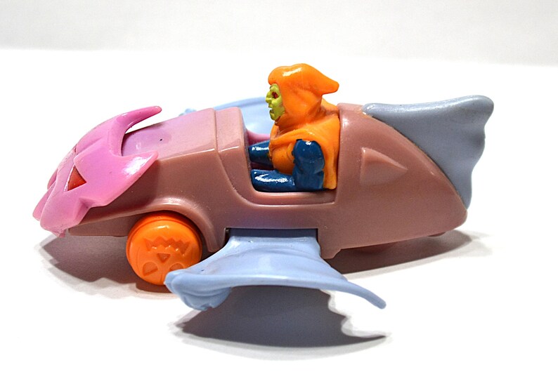 May include: A vintage toy vehicle featuring a character figure. The vehicle is primarily brown with pink and blue accents, and orange wheels. The character has an orange hood and green skin. The toy is on a white surface.