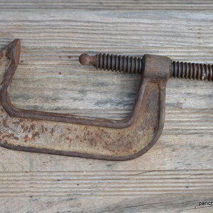 May include: A rusty metal C-clamp with a threaded screw and a wing nut. The clamp is on a wooden surface.