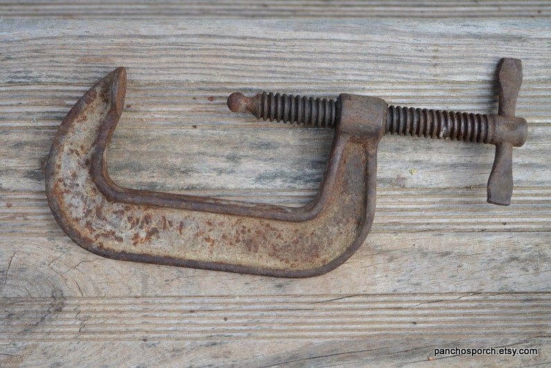 May include: A rusty metal C-clamp with a threaded screw and a wing nut. The clamp is on a wooden surface.