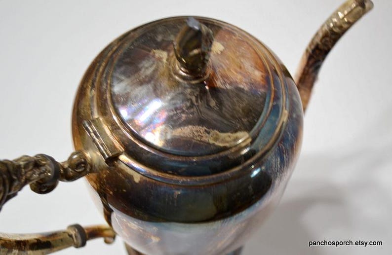 Pu&ograve; includere: Una teiera argentata con un manico e un coperchio decorativi. La teiera ha una superficie lucida e riflettente con una finitura leggermente iridescente.