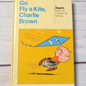 May include: A vintage paperback book titled "Go Fly a Kite, Charlie Brown" by Charles M. Schulz. The cover features a cartoon illustration of Charlie Brown running with a kite against a yellow background.