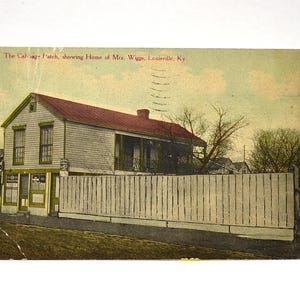 May include: A vintage postcard featuring a two-story house with a red roof and light-colored siding, enclosed by a tall white wooden fence. The text at the top reads: The Cabbage Patch, showing Home of Mrs. Wiggs, Louisville, Ky. A tree stands nearby.