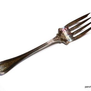 May include: An antique silver fork with four tines, a decorative floral design on the handle, and a slightly tarnished finish. The fork is set against a white background, showcasing its vintage charm.