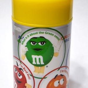 May include: A cylindrical container with a yellow lid featuring M&M's characters. The container has a silver body with colorful graphics of the candies, including the text "What is it about the Green ones?" and "I'm Red Hot!"