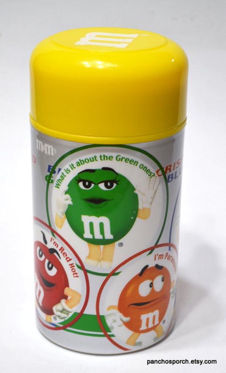 May include: A cylindrical container with a yellow lid featuring M&M's characters. The container has a silver body with colorful graphics of the candies, including the text "What is it about the Green ones?" and "I'm Red Hot!"