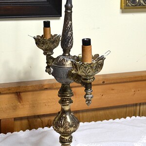 Puede incluir: Una l&aacute;mpara de mesa de dos luces estilo candelabro con acabado met&aacute;lico en tonos dorados y gris oscuro. La l&aacute;mpara presenta una base y un tallo central texturizados y ornamentados con detalles de hojas, dos casquillos y un asa superior.