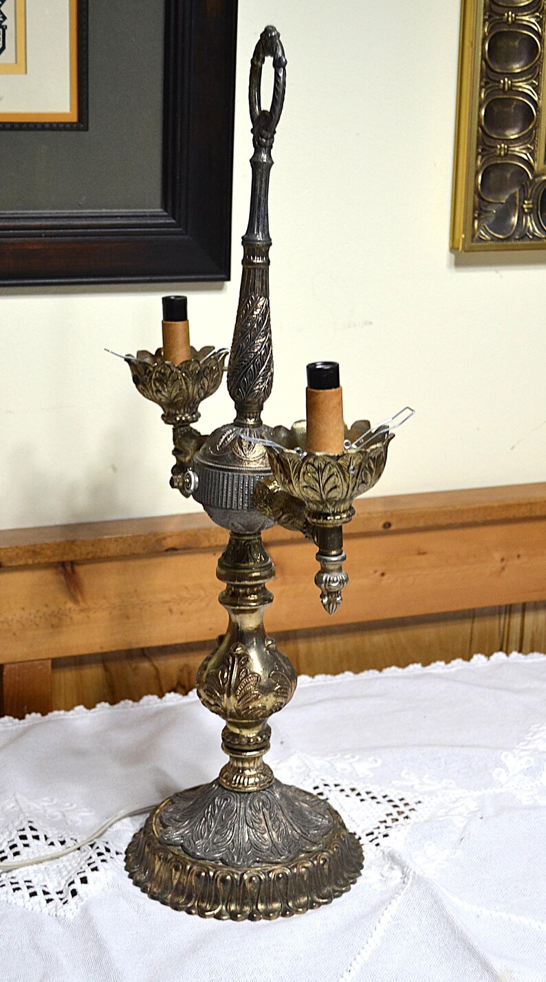 Puede incluir: Una l&aacute;mpara de mesa de dos luces estilo candelabro con acabado met&aacute;lico en tonos dorados y gris oscuro. La l&aacute;mpara presenta una base y un tallo central texturizados y ornamentados con detalles de hojas, dos casquillos y un asa superior.