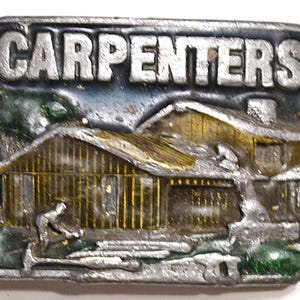 May include: A rectangular metal belt buckle with the word "CARPENTERS" in raised silver letters. The buckle features a detailed relief of a wooden house with a carpenter working outside. The background has blue and green accents.