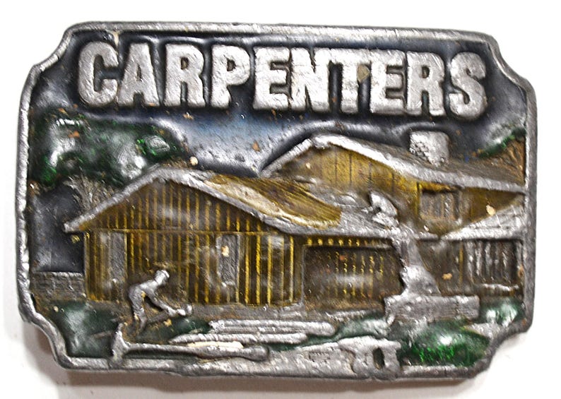 May include: A rectangular metal belt buckle with the word "CARPENTERS" in raised silver letters. The buckle features a detailed relief of a wooden house with a carpenter working outside. The background has blue and green accents.