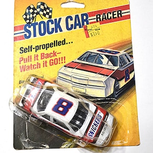 May include: A vintage toy stock car racer on its original packaging. The car is white with red and blue accents, featuring the number 8 and "Snickers" branding. The packaging reads "Stock Car Racer" and "Self-propelled... Pull it Back - Watch it GO!!!"
