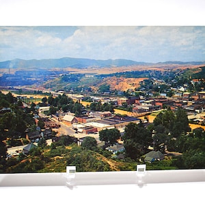 May include: Vintage postcard featuring a scenic aerial view of a town nestled in a valley, surrounded by rolling hills and mountains under a cloudy sky. The town includes buildings, trees, and a river.