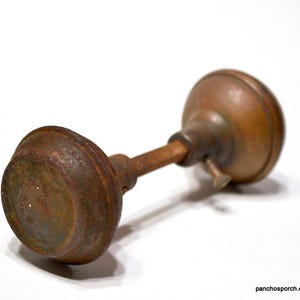 May include: An antique door knob with a weathered, rusty finish. The knob has a rounded shape with a cylindrical connecting rod. The metal has a mix of brown and green hues, suggesting age and exposure to the elements.