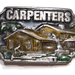 May include: A rectangular metal belt buckle with the word "CARPENTERS" in raised letters. The buckle features a detailed scene of a wooden house with a carpenter working outside. The buckle has a silver finish with touches of green and gold.