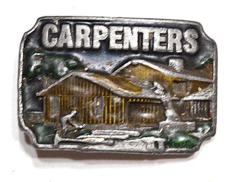 May include: A rectangular metal belt buckle with the word "CARPENTERS" in raised letters. The buckle features a detailed scene of a wooden house with a carpenter working outside. The buckle has a silver finish with touches of green and gold.