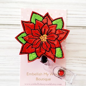 May include: Red poinsettia flower embroidered on a white background with green leaves. The flower is attached to a clear plastic badge holder with a red jewel.