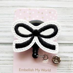 Puede incluir: Lazo blanco y negro con un carrete retr&aacute;ctil de credencial negro. El carrete de credencial est&aacute; unido a una tarjeta rosa con lunares blancos y el texto "Embellish My World Boutique".