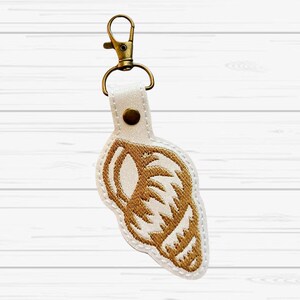 May include: A white seashell-shaped keychain with gold embroidery and a gold-colored clasp. The keychain is attached to a white strap with a gold rivet. The design features a detailed embroidered seashell.