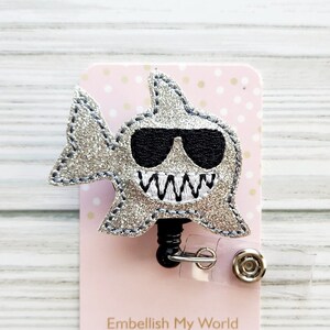 May include: Silver glitter shark with black sunglasses and a white stitched smile. The shark is attached to a retractable badge reel with a clear plastic strap and a silver button.