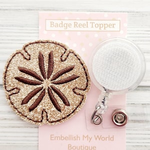 May include: A gold glitter sand dollar badge reel topper with a brown embroidered design. The badge reel is clear with a white fabric backing. The card reads "Badge Reel Topper" and "Embellish My World Boutique".