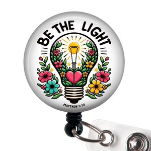 May include: A white retractable badge holder with a round, colorful lightbulb design. The lightbulb is surrounded by flowers and the text "Be the Light" with the scripture reference "Matthew 5:14".