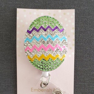 K&ouml;nnte beinhalten: Ein gr&uuml;ner, lila, blauer, rosa und gelber Oster-Eier-f&ouml;rmiger Badgehalter mit Strasssteinen. Der Badgehalter ist an einer durchsichtigen, einziehbaren Badge-Rolle aus Kunststoff befestigt. Der Badgehalter befindet sich auf einer rosa Karte mit wei&szlig;en Punkten. Die Karte tr&auml;gt den Schriftzug "Embellish My World Boutique" und "www.embellishmyworld.etsy.com".