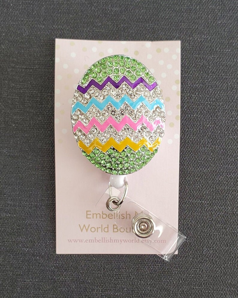 K&ouml;nnte beinhalten: Ein gr&uuml;ner, lila, blauer, rosa und gelber Oster-Eier-f&ouml;rmiger Badgehalter mit Strasssteinen. Der Badgehalter ist an einer durchsichtigen, einziehbaren Badge-Rolle aus Kunststoff befestigt. Der Badgehalter befindet sich auf einer rosa Karte mit wei&szlig;en Punkten. Die Karte tr&auml;gt den Schriftzug "Embellish My World Boutique" und "www.embellishmyworld.etsy.com".