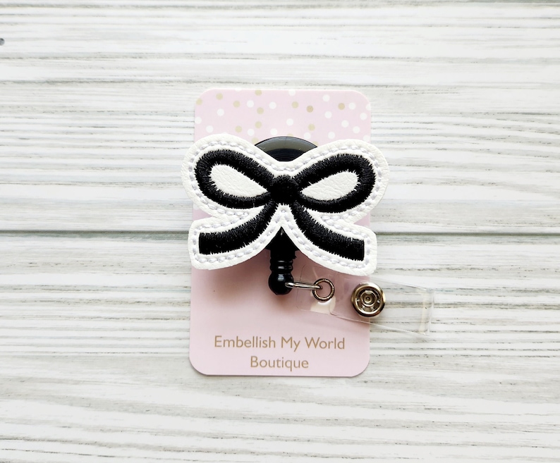 Puede incluir: Lazo blanco y negro con un carrete retr&aacute;ctil de credencial negro. El carrete de credencial est&aacute; unido a una tarjeta rosa con lunares blancos y el texto "Embellish My World Boutique".