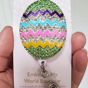 K&ouml;nnte beinhalten: Ein gr&uuml;n, gelb, rosa, blau und violetter Oster-Eier-f&ouml;rmiger Badge-Halter mit Strasssteinen. Der Badge-Halter ist an einer durchsichtigen, einziehbaren Badge-Rolle mit einem silbernen Drehclip befestigt. Der Text "Embellish My World Boutique" und "www.embellishmyworld.com" ist auf die wei&szlig;e Karte hinter dem Badge-Halter gedruckt.
