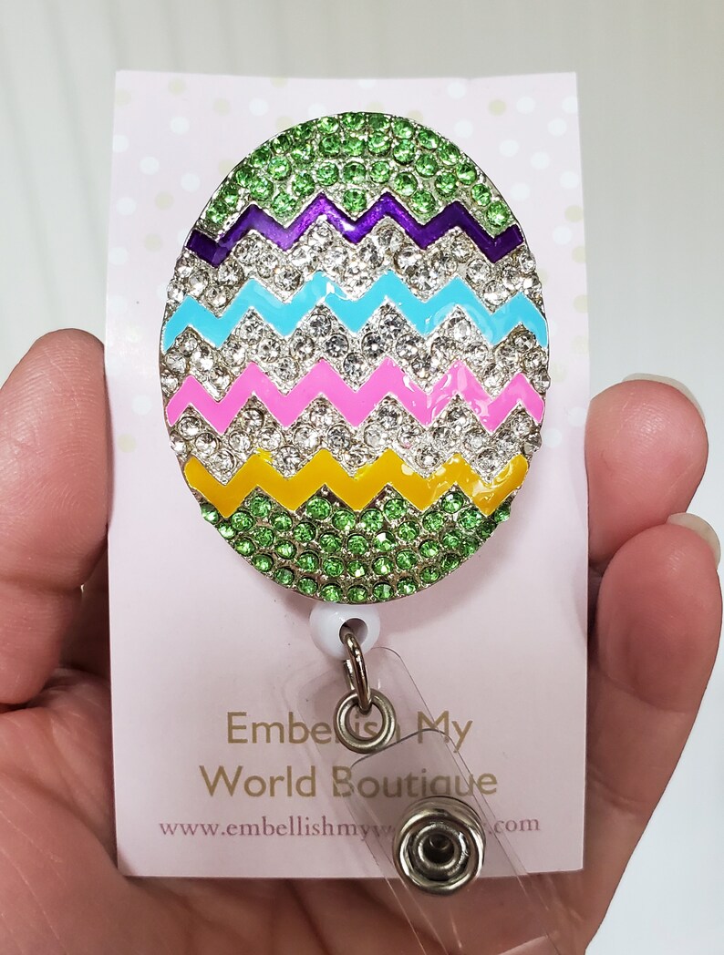 K&ouml;nnte beinhalten: Ein gr&uuml;n, gelb, rosa, blau und violetter Oster-Eier-f&ouml;rmiger Badge-Halter mit Strasssteinen. Der Badge-Halter ist an einer durchsichtigen, einziehbaren Badge-Rolle mit einem silbernen Drehclip befestigt. Der Text "Embellish My World Boutique" und "www.embellishmyworld.com" ist auf die wei&szlig;e Karte hinter dem Badge-Halter gedruckt.