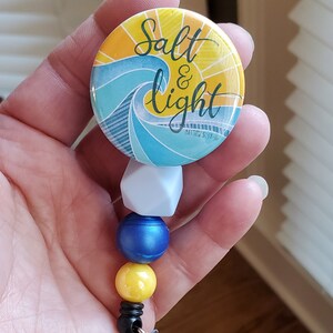 May include: A beaded retractable badge holder with a blue, yellow, and white color scheme. The badge holder features a round button with a wave design and the text "Salt & Light Matthew 5:13-16".