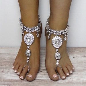 May include: Silver-toned foot jewellery with clear, sparkling rhinestones. The anklets have a large, round centrepiece, a teardrop pendant, and a chain connecting to the toe. The jewellery is displayed on a light wood surface.
