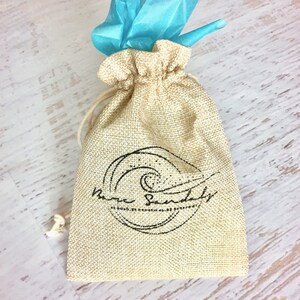 Peut inclure: Un sac en toile de jute beige avec cordon de serrage portant l'inscription "Bare Sandals" en noir. Le sac est nou&eacute; avec une ficelle beige et surmont&eacute; d'un paquet de papier de soie bleu vif. Le sac est pos&eacute; sur une surface en bois clair.