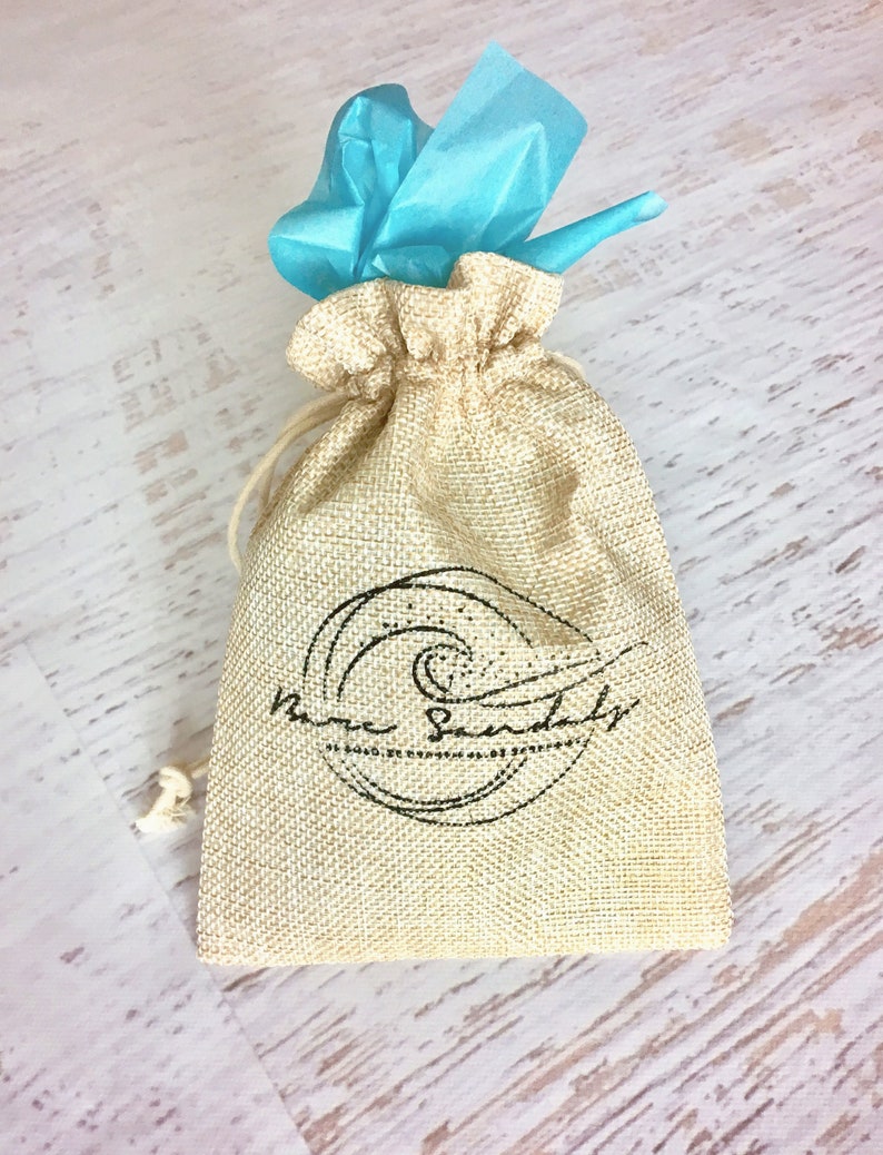 Peut inclure: Un sac en toile de jute beige avec cordon de serrage portant l'inscription "Bare Sandals" en noir. Le sac est nou&eacute; avec une ficelle beige et surmont&eacute; d'un paquet de papier de soie bleu vif. Le sac est pos&eacute; sur une surface en bois clair.