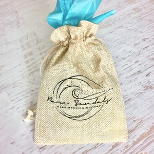May include: A small, beige burlap bag with a drawstring closure. The bag has a black logo with a wave design and the text "The Soundly". The bag is tied with a light blue tissue paper.