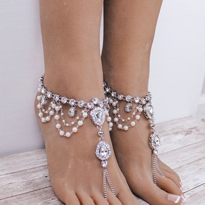 Puede incluir: Un par de sandalias descalzas plateadas con adornos de cristal y detalles de perlas. Las sandalias tienen un delicado dise&ntilde;o de cadena y un colgante de cristal.