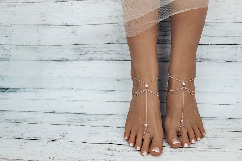 May include: A pair of delicate gold foot chains with pearl accents. The chains are layered and feature a delicate design. The chains are worn on a person's feet, with the ends of the chains extending to the toes.