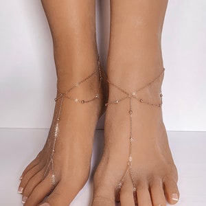 Peut inclure: Gros plan sur des pieds orn&eacute;s de sandales dor&eacute;es. Les sandales sont compos&eacute;es d'une cha&icirc;ne avec de petites perles et des d&eacute;tails en forme d'&eacute;toiles, reliant la cheville et s'&eacute;tendant jusqu'aux orteils. Les ongles sont peints avec une manucure fran&ccedil;aise.